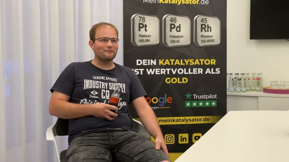 Mann sitzt vor Banner mit Text "DEIN KATALYSATOR IST WERTVOLLER ALS GOLD" und Periodensystem-Elementen.