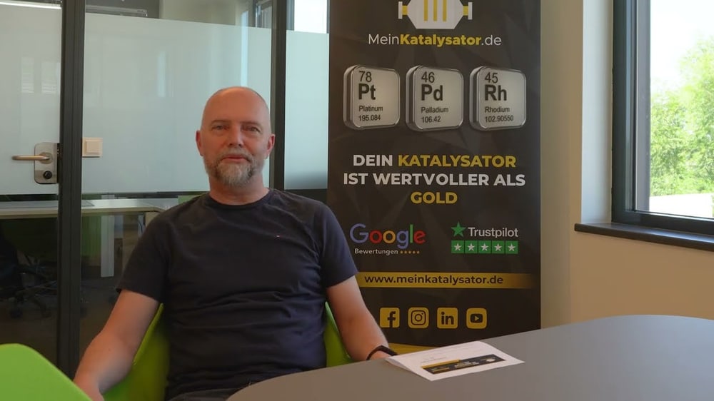 Mann sitzt vor einem Banner mit "DEIN KATALYSATOR IST WERTVOLLER ALS GOLD" und Logos.